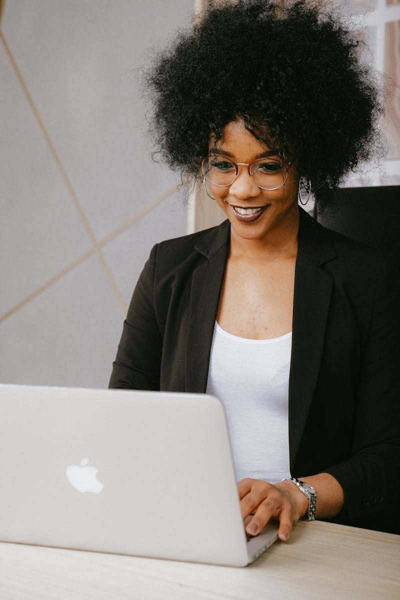 pexels-photo-3727458-3727458 Smiling woman with afro hair working on a laptop in a modern office setting.